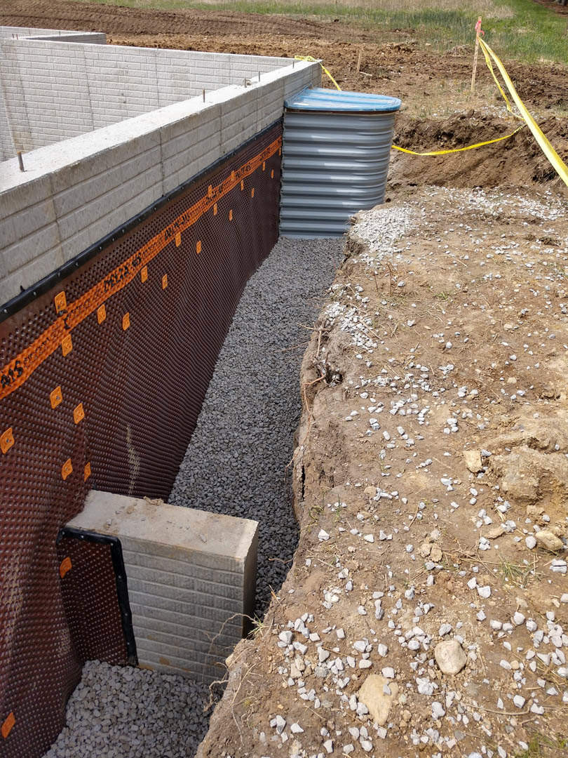 Concrete foundation with exposed rebar, dirt and gravel surrounding the base, metal barrel with blue lid, nearby fence, scattered rocks and concrete blocks, patches of grass and