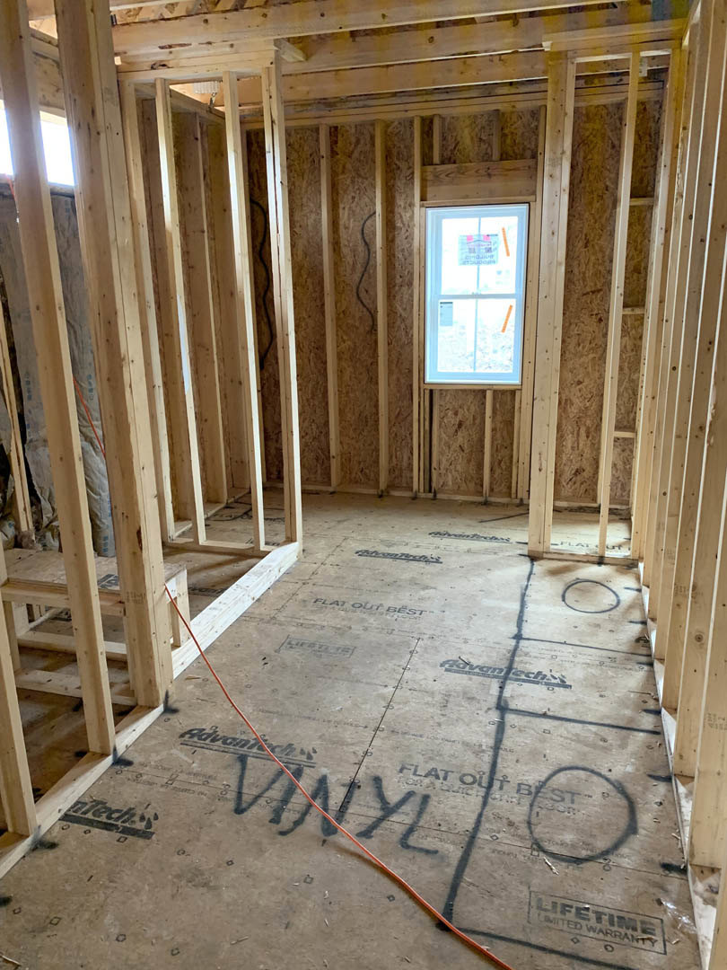 Wood-framed room under construction with exposed beams, unfinished floor marked with black writing, and a window displaying a sign.