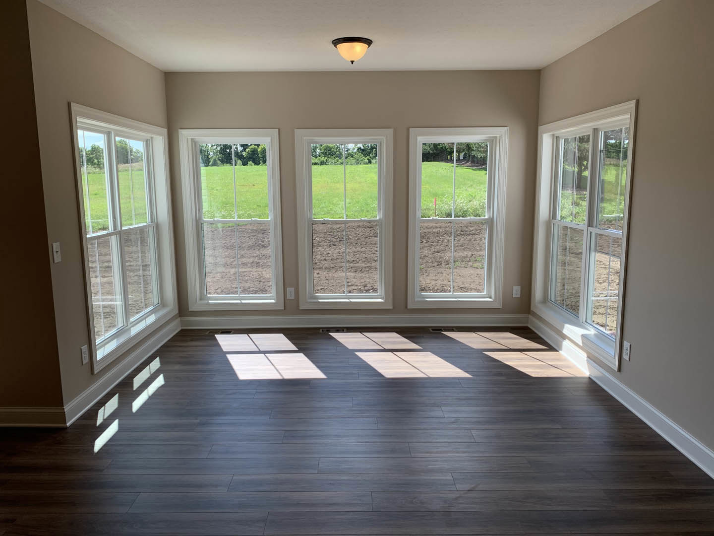 Spacious room featuring wood flooring with white square inlays, large windows framed in white overlooking a green field, neutral walls, and a ceiling-mounted light fixture with a