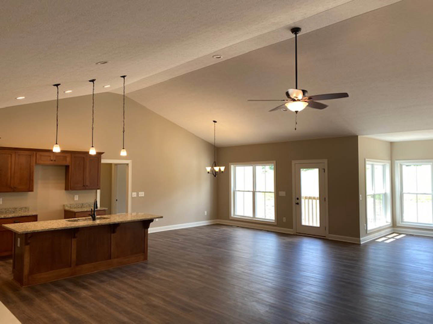 Spacious open-concept kitchen and living area featuring a central island with sink, wood flooring, ceiling fan with light fixture, white door with window, and sunlight streaming
