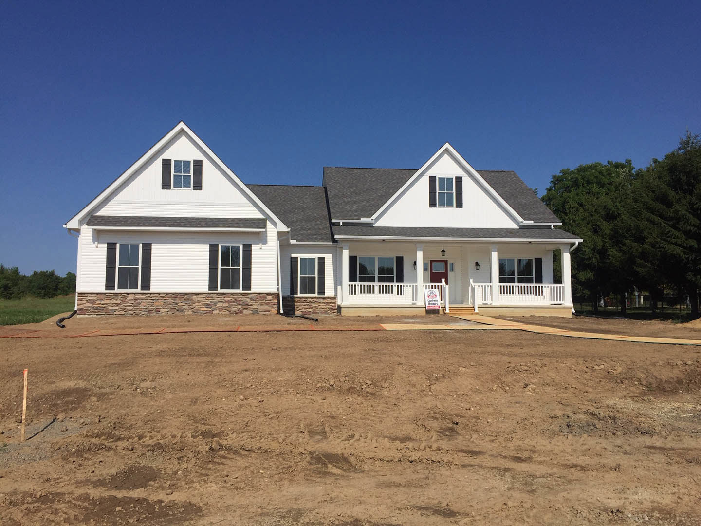 White house with stone and brick lower walls, white porch railing, and white-framed windows, set beside a dirt area with tire tracks under a clear blue sky