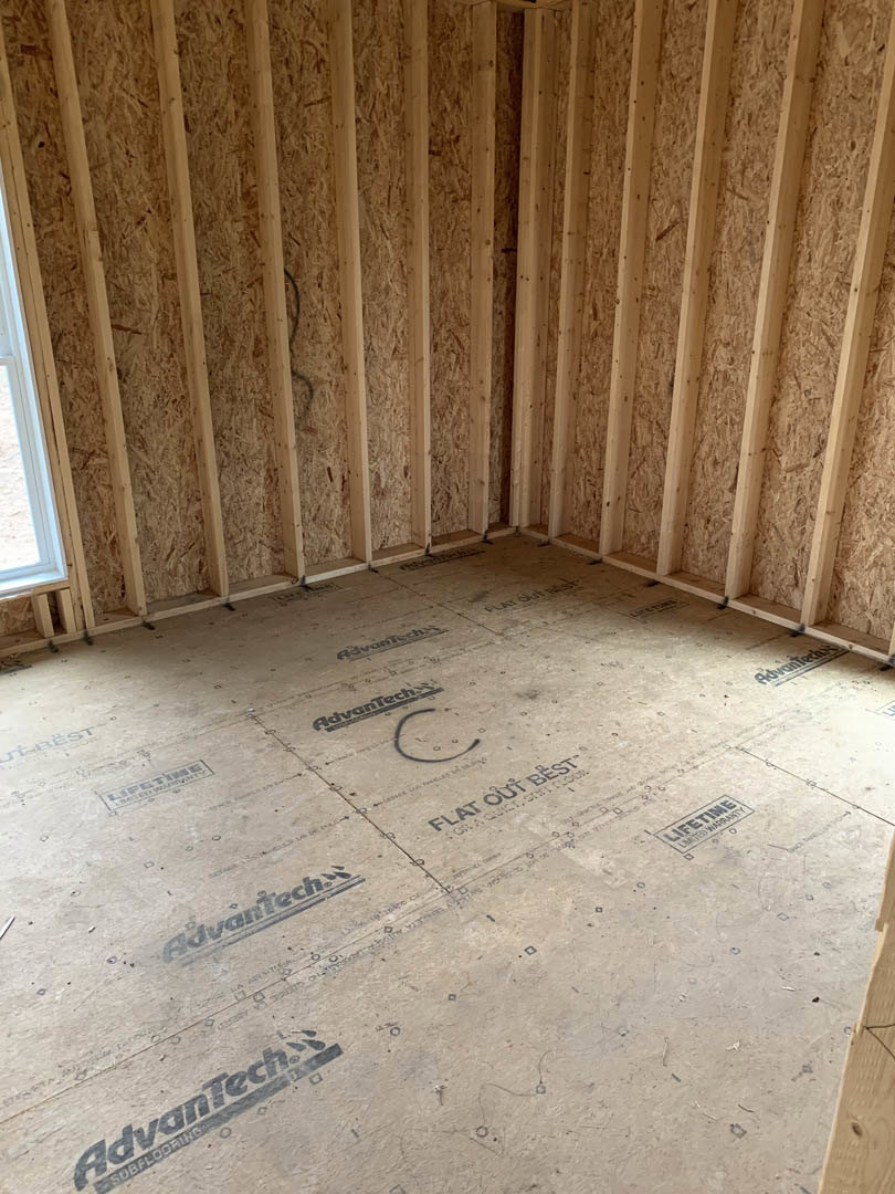 Wood-framed room under construction with exposed beams, unfinished floor, and handwritten logo on a white surface; black wire visible against plaster wall.