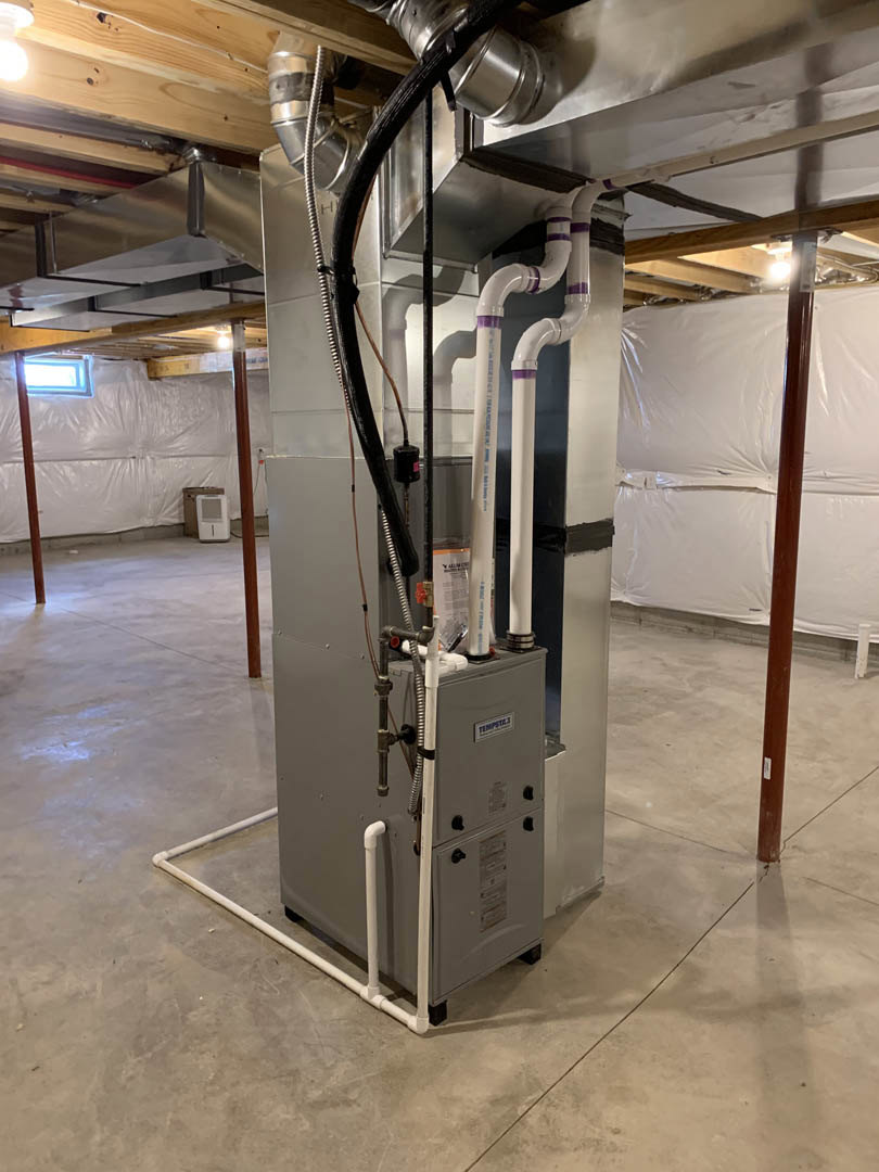 Mechanical room with exposed steel pipes, hoses, and wires connected to a large machine; white plastic sheeting covers part of the wall and floor.