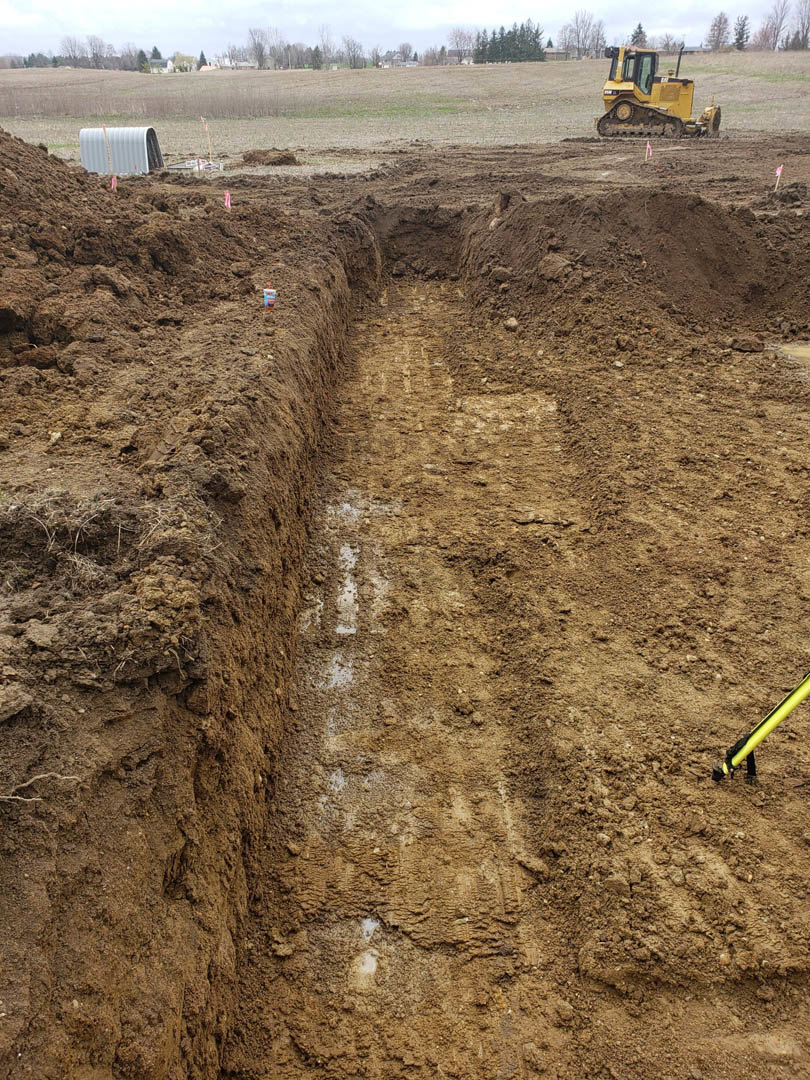 Wide trench dug in brown dirt field with yellow bulldozer parked nearby, white metal tunnel structure partially visible, yellow pole close-up in foreground, exposed dirt wall, and