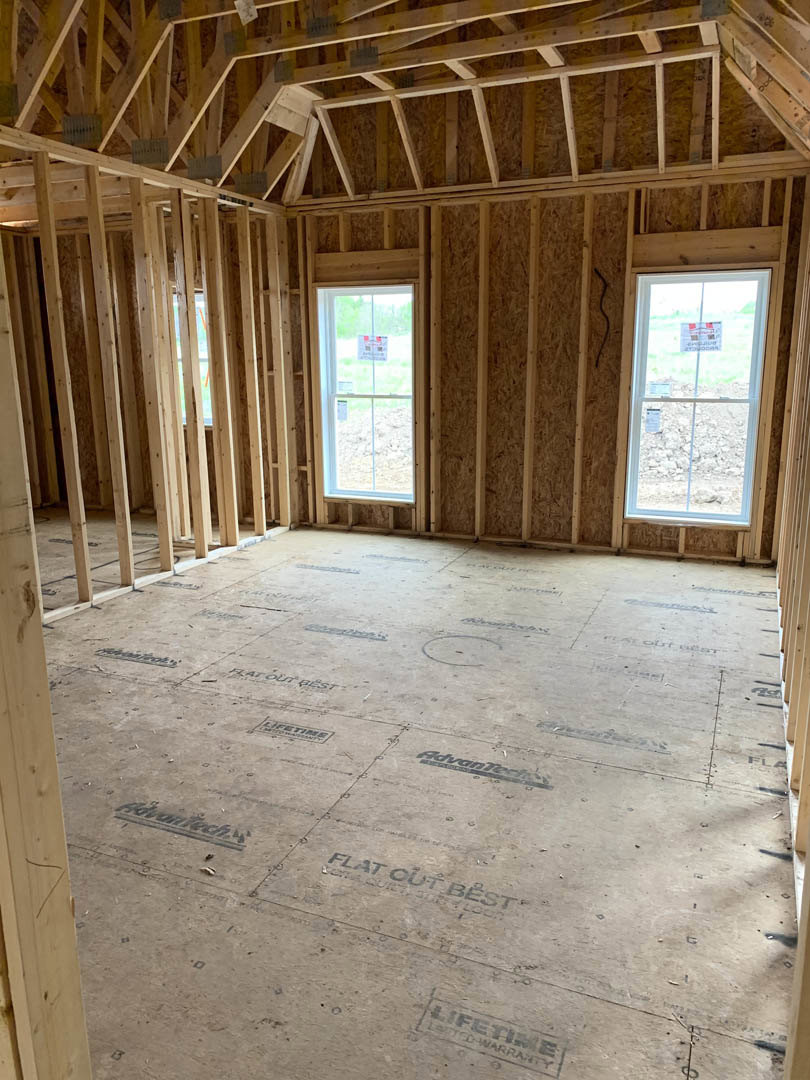 Room under construction with exposed wood framing, ceiling trusses, multiple windows, and a floor marked with text and wood beams; window features a visible sign.