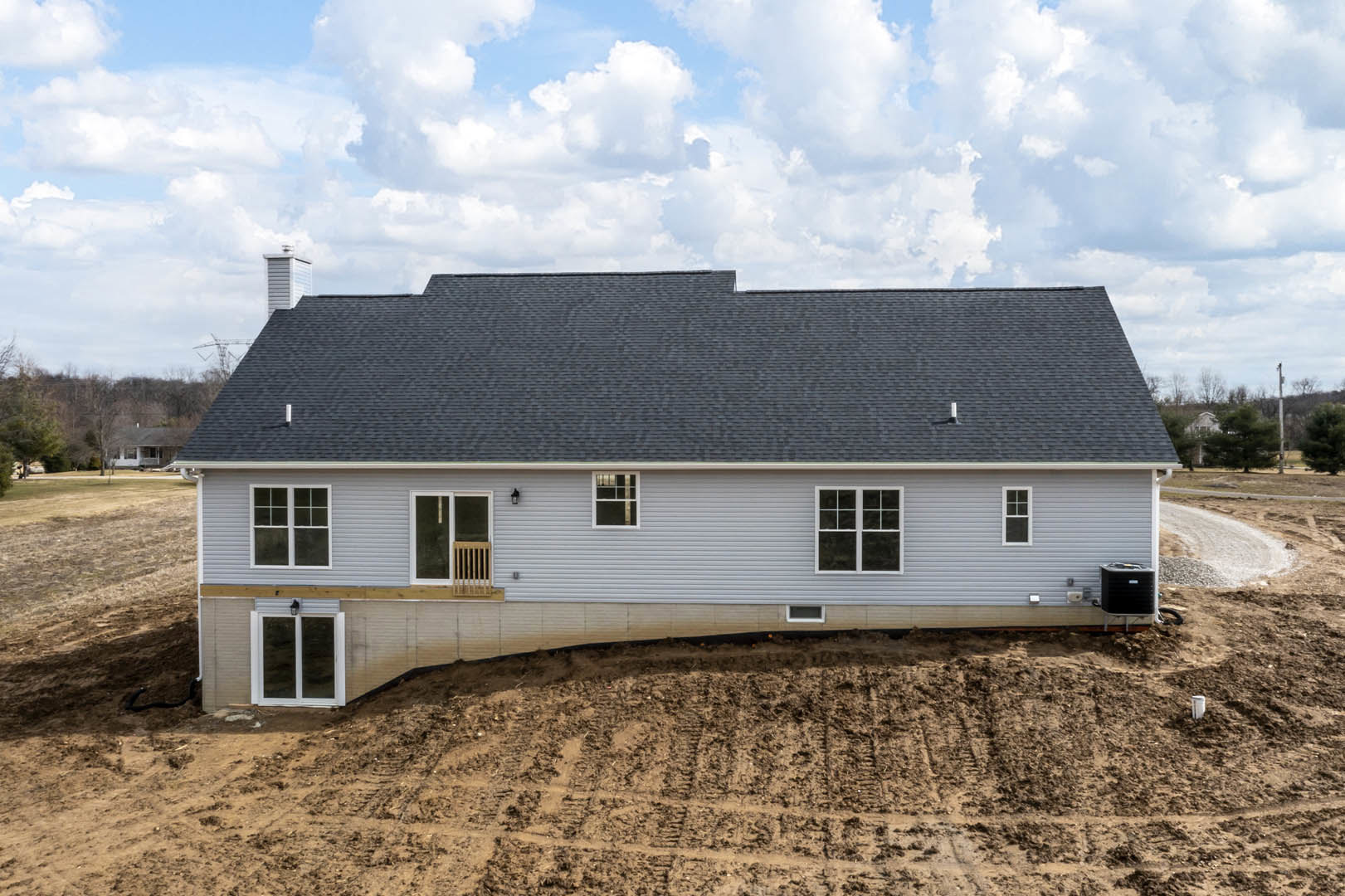 Partially built house with exposed wooden framing, white-framed windows, unfinished roof, and surrounding dirt field under cloudy sky