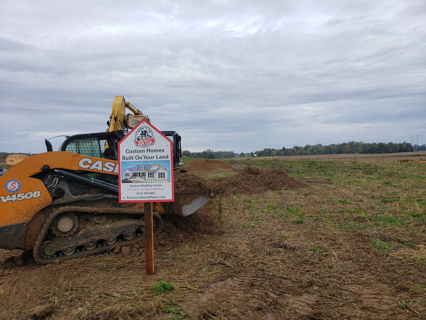 Bulldozer parked on a dirt field with grass and clouds in the background, construction sign mounted on a post nearby