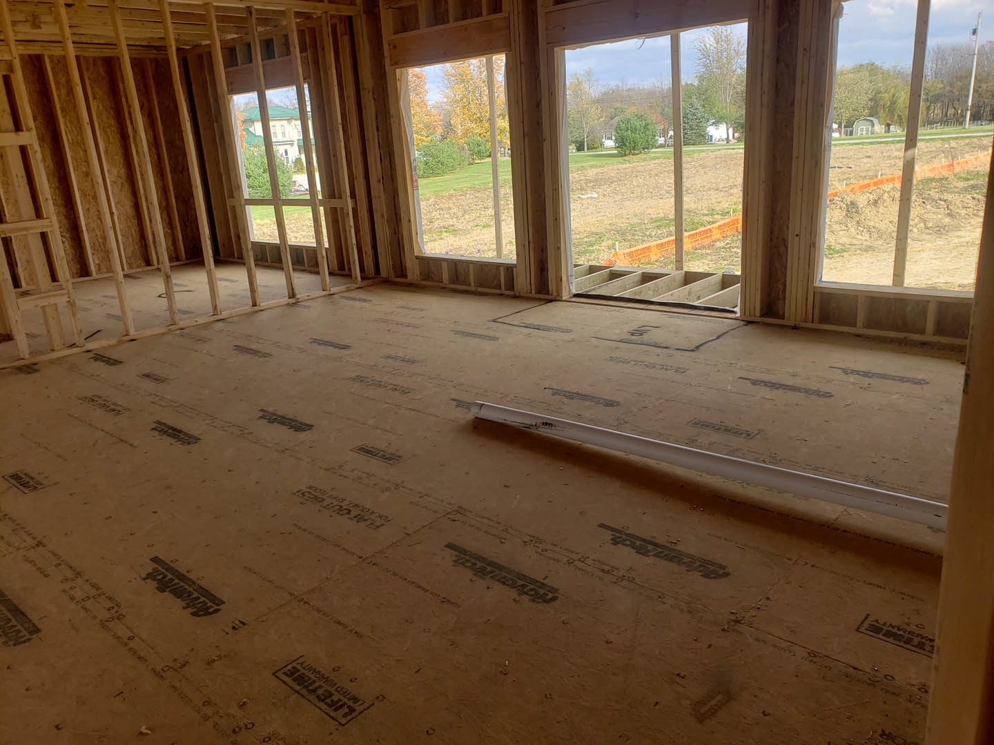 Wood-framed room with large windows, sandy floor showing tire tracks, white pipe along wall, and leafy trees visible outside