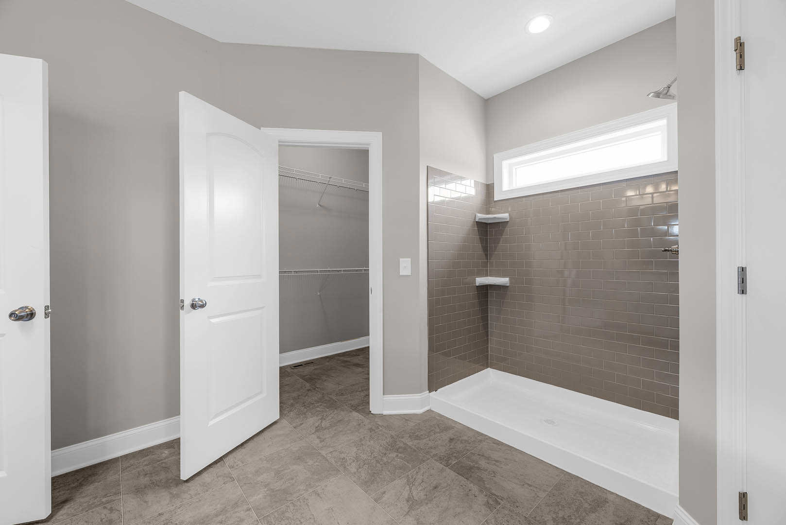 Bathroom with brown tile floor, white shower base, open white door with silver handle, light fixture above tiled wall