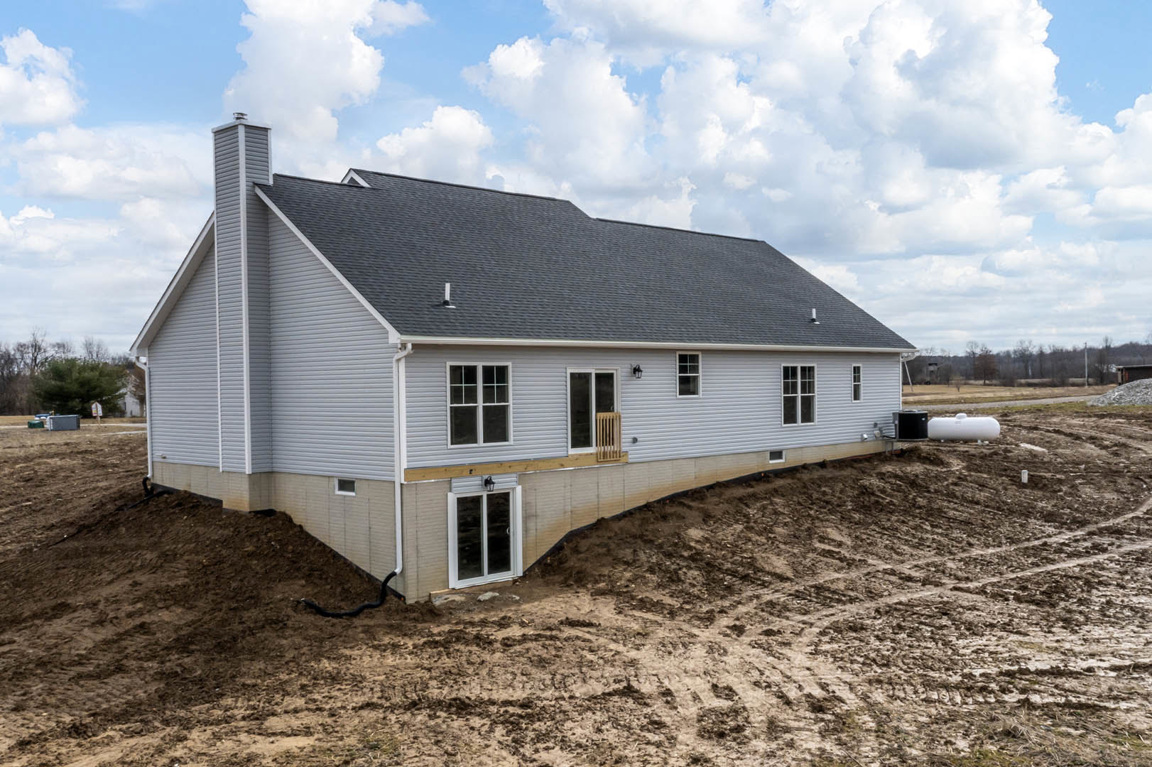 Two-story custom home with stone foundation, white siding, large windows, wooden railing, and shingled roof beneath a cloudy sky; construction materials visible in foreground, hill
