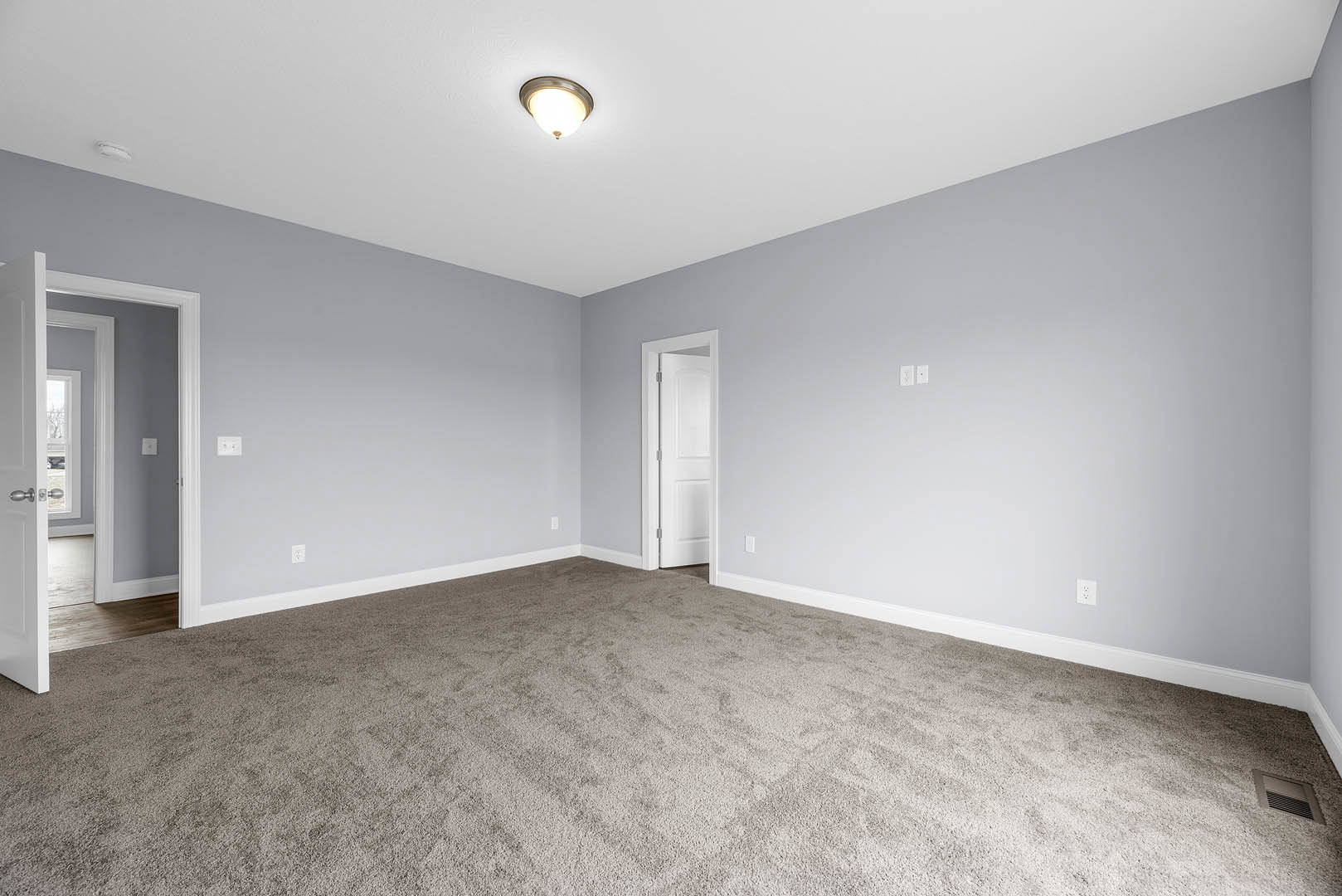 Neutral-toned carpeted room with white walls, open white door with metal frame, ceiling-mounted light fixture, and simple molding along the baseboards