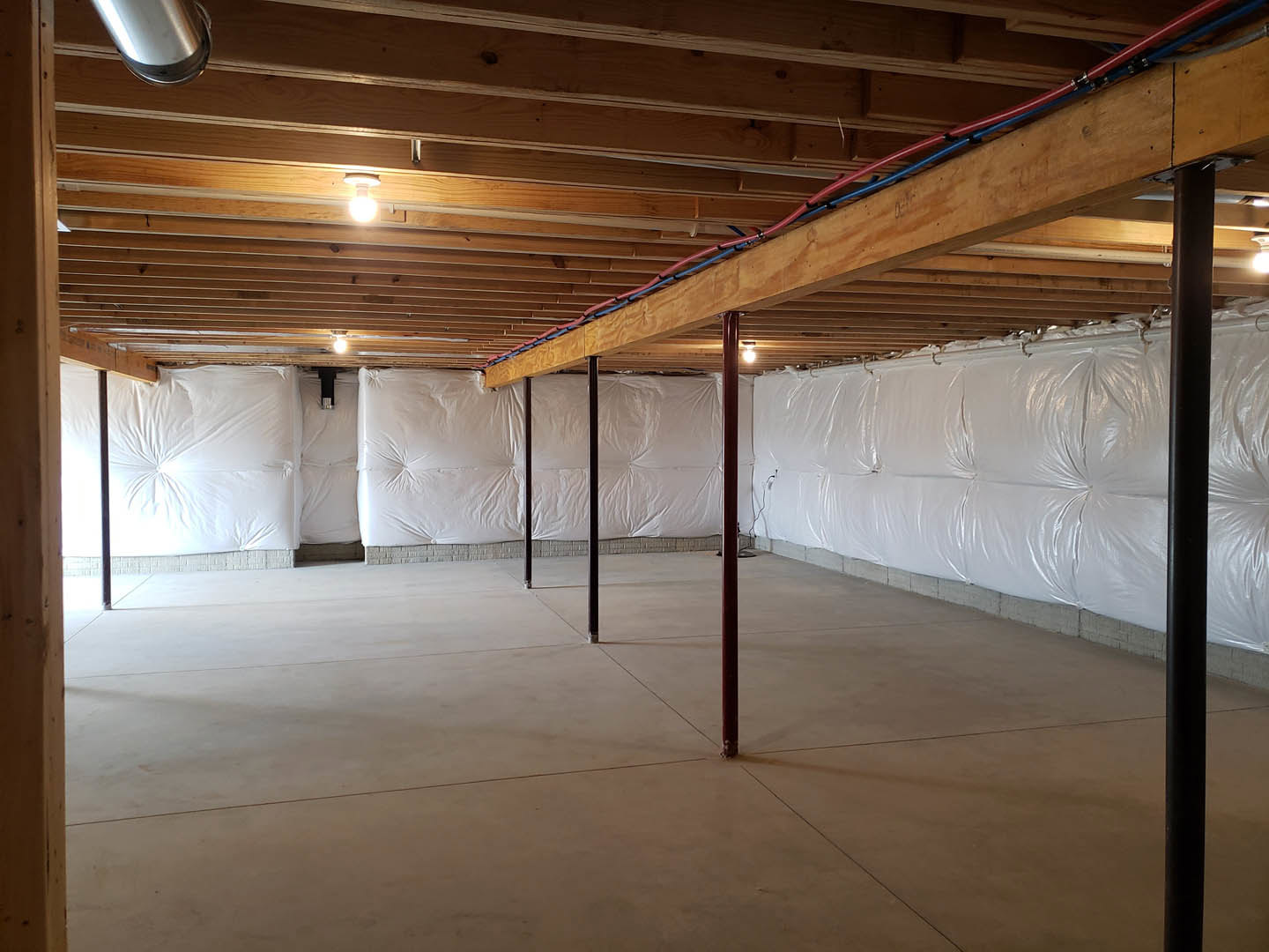 Unfinished room with exposed wooden ceiling beam, white plastic sheeting covering walls and floor, visible metal pipe, and white mattress on floor