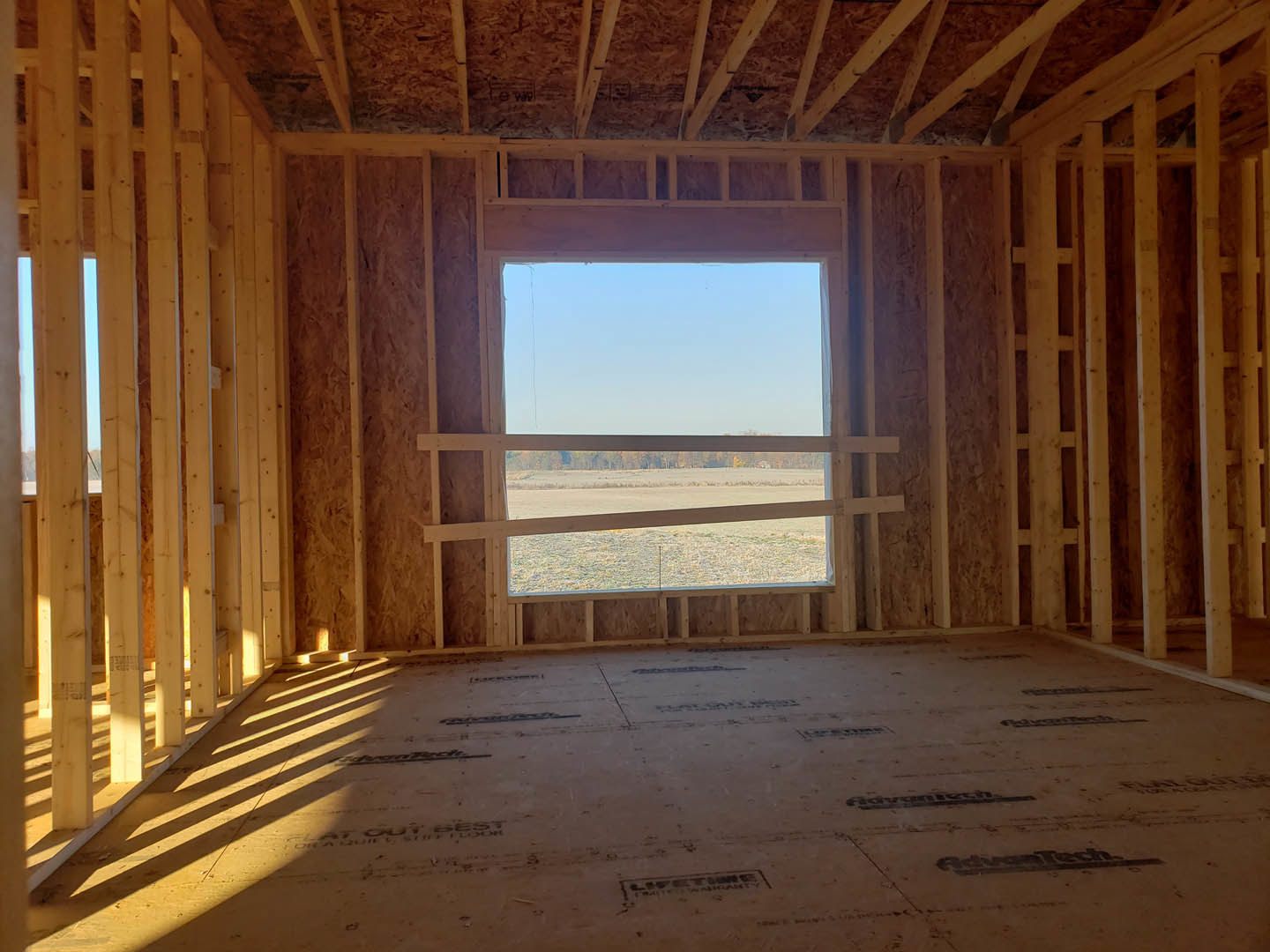 Sunlit room with exposed wood ceiling beams, large window framing blue sky and open field view, light-colored walls, and hardwood flooring