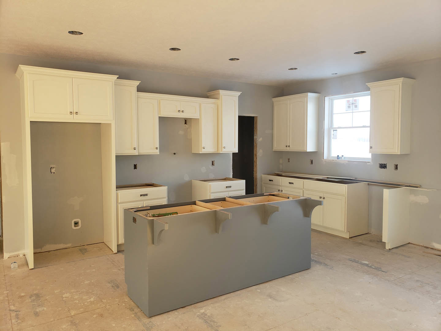 White kitchen cabinets, gray island countertop, four open shelves, window with newspaper, wall-mounted electrical outlets, recessed ceiling lights, light wood flooring, stainless