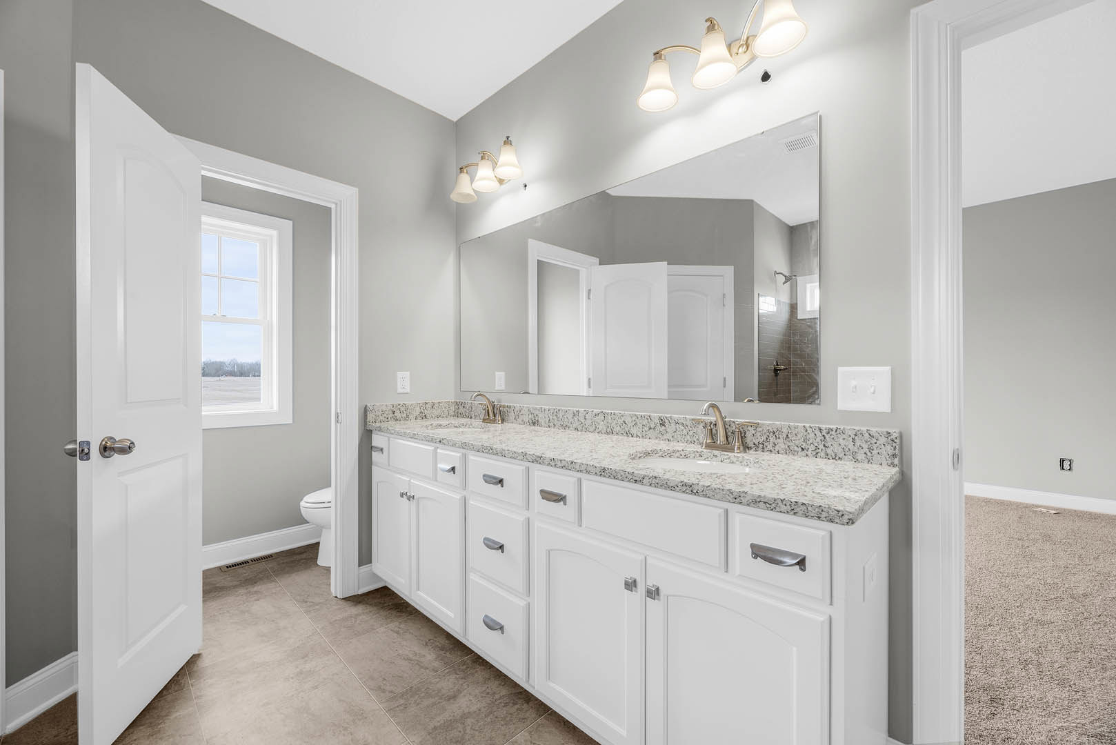 Bathroom with white cabinets, wide mirror above sink, three-light fixture, tile flooring, window showing snowy landscape, open white door leading to adjacent room