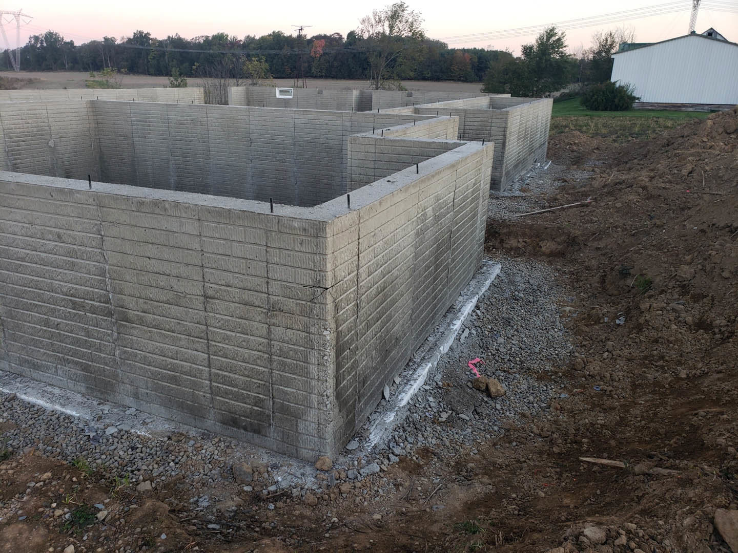 Concrete block wall with square opening set in grassy field, white metal building with sloped roof nearby, power lines and dense forest in background, pink paper resting on rocks