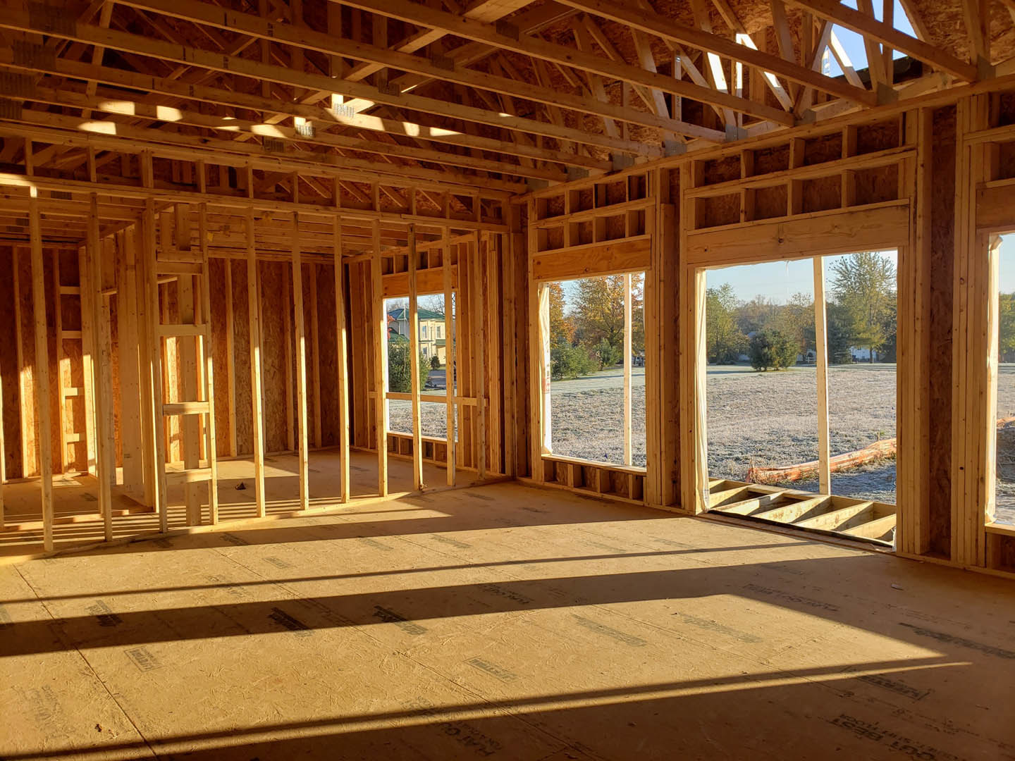 Open room with exposed wood ceiling beams, large windows overlooking grassy field and leafy trees, unfinished wooden floor, construction materials visible