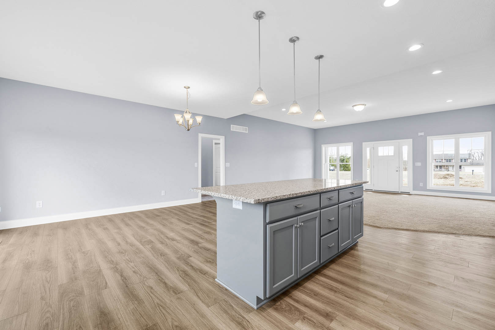 Spacious kitchen featuring a large granite island, white cabinetry, stainless steel sink, pendant chandelier, and a window overlooking a grassy field.