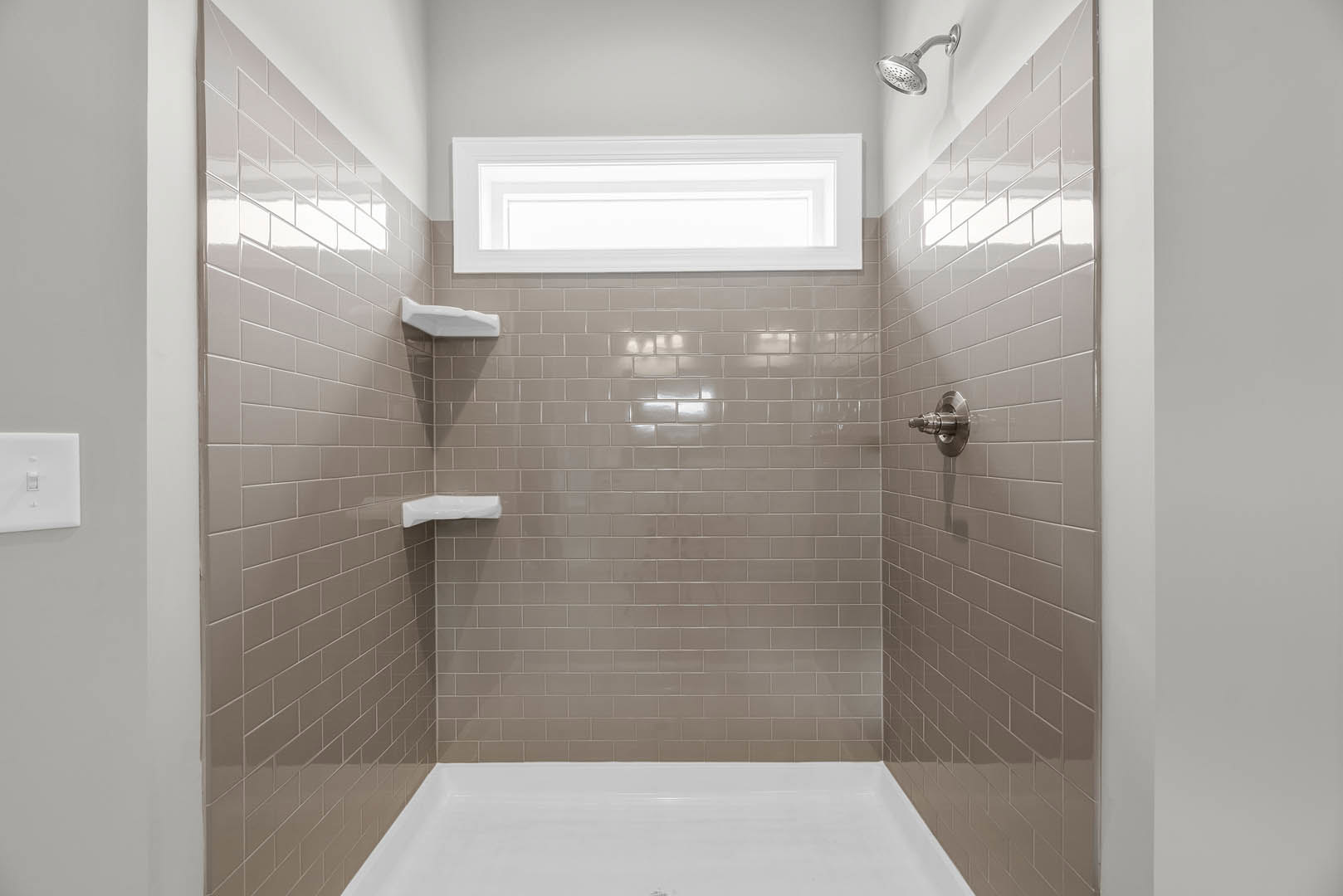 Walk-in shower with white tile walls, chrome shower head, and frosted window above; partial view of white bathtub and toilet nearby
