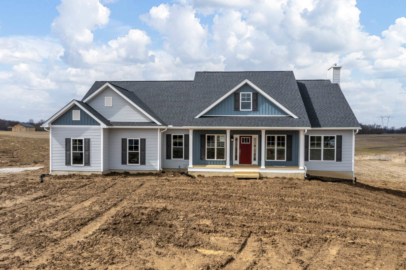 Two-story house with white siding, red front door, covered porch, white-framed windows, and dirt yard under blue sky with scattered clouds
