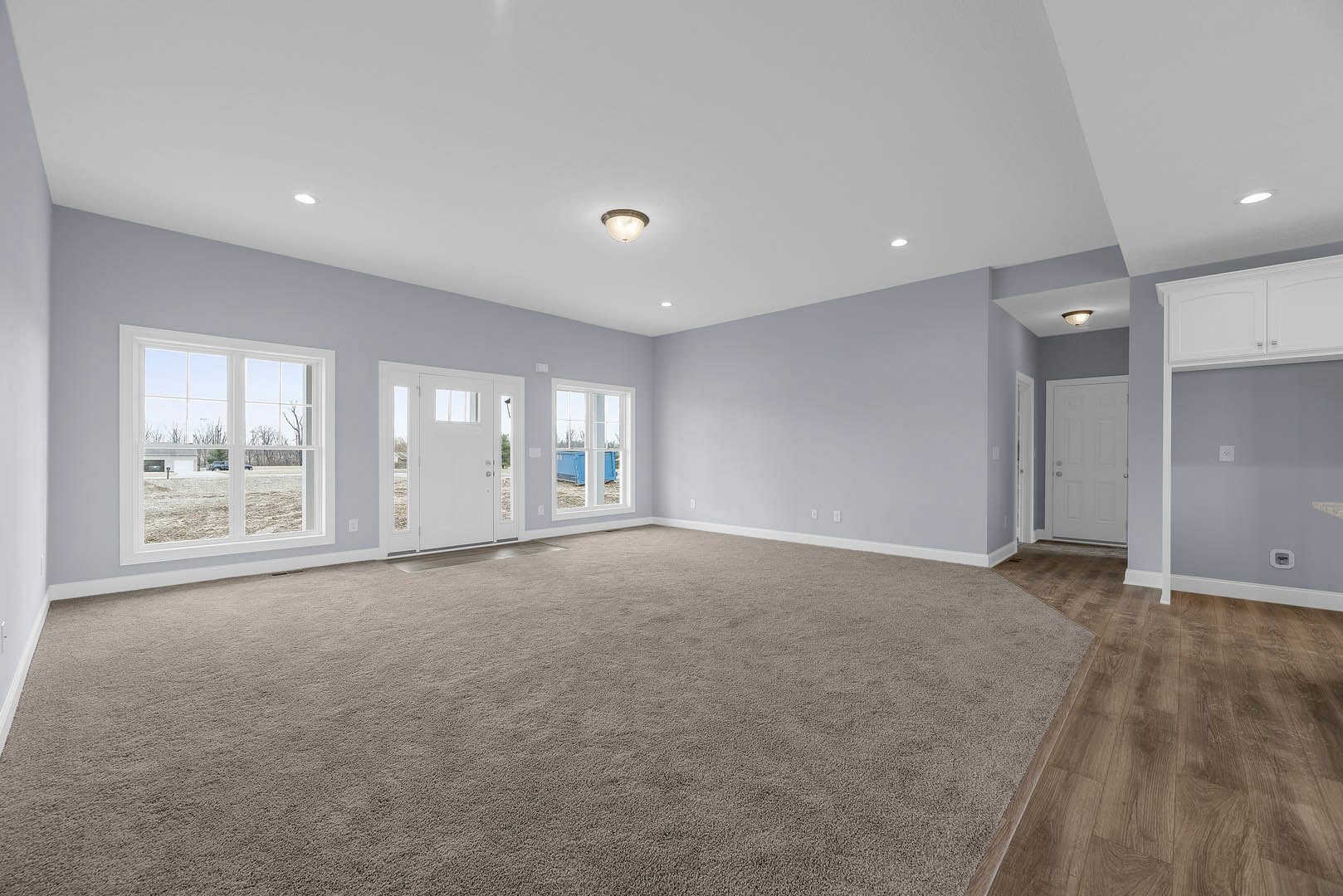 White door with silver knobs, gray carpeted floor, large window overlooking a grassy field, white walls, ceiling-mounted light fixture