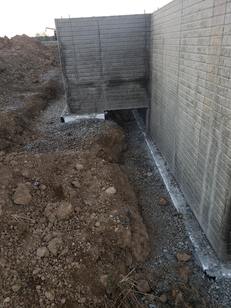 Red brick exterior wall with a hole in the ground surrounded by dirt, gravel, and concrete curb