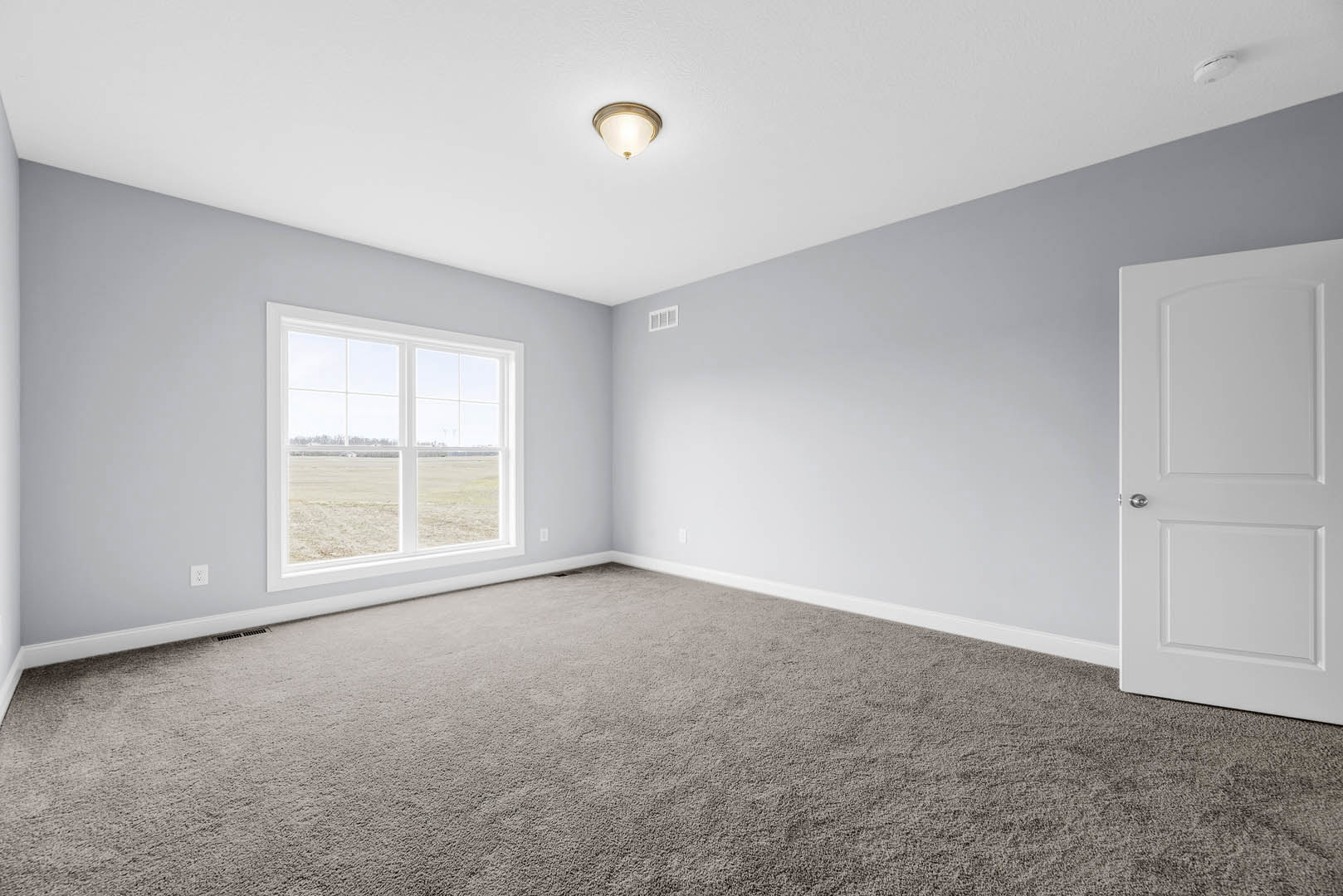 Carpeted room with white walls, large window overlooking grassy field, white door, ceiling-mounted light fixture and smoke detector