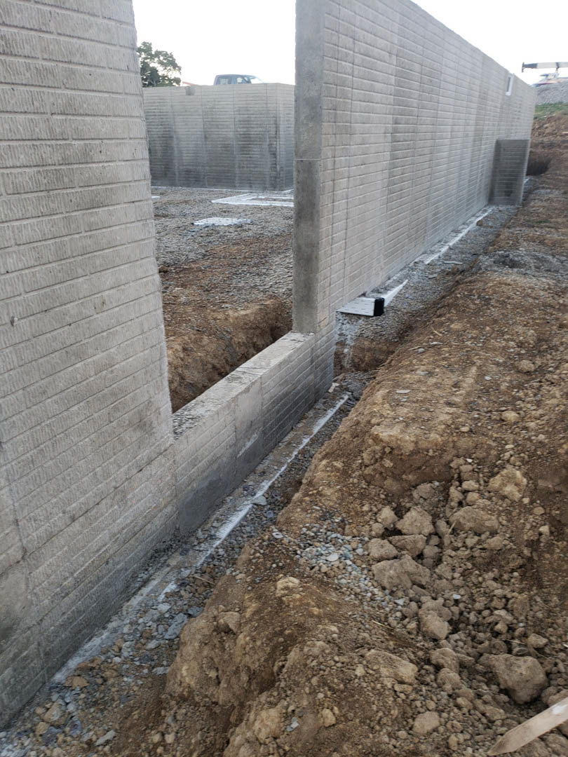 Concrete foundation wall with exposed soil and excavation hole, adjacent to white and grey brick surfaces, leafy tree visible in background.