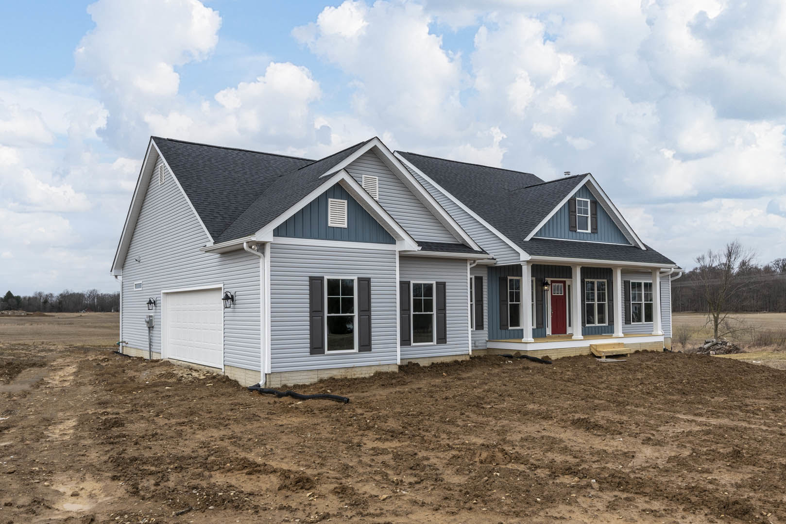Two-story house under construction with white-framed windows, exposed dirt yard, and construction pipe; neighboring homes visible under partly cloudy blue sky.