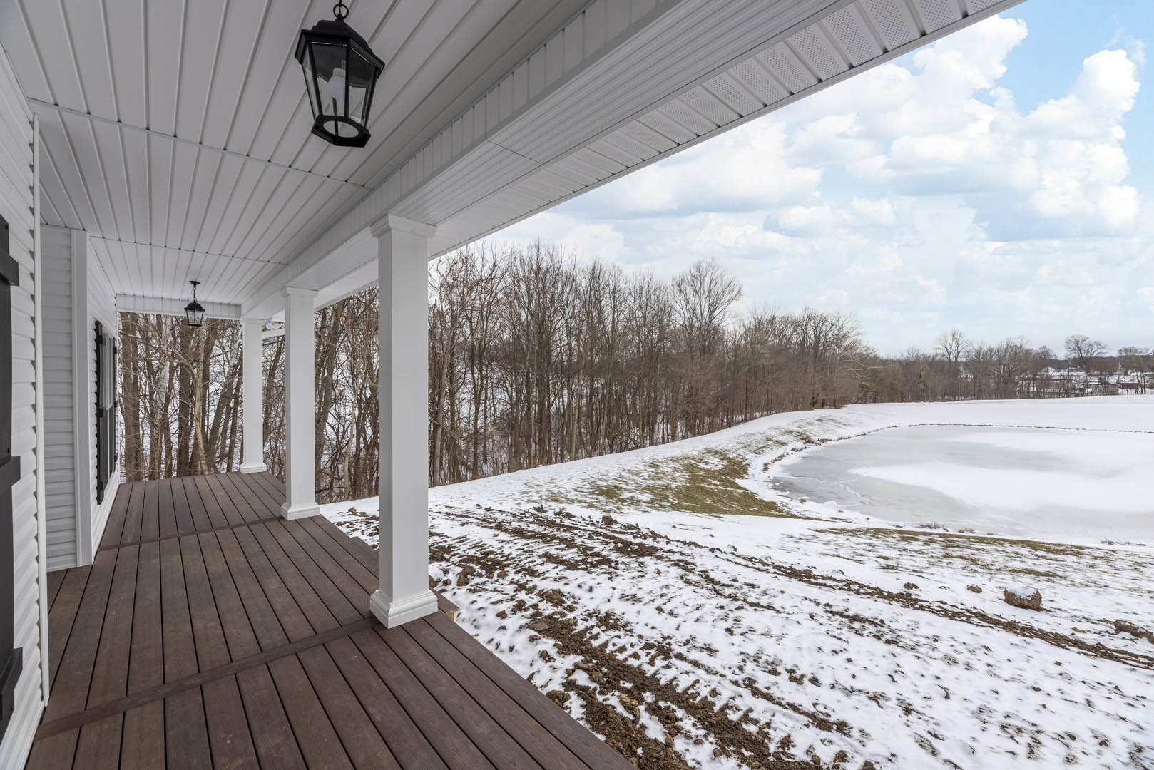 Covered porch with white pillars, black and white sign, wall-mounted light fixture, snow-covered deck, snowy field, lake, and leafless trees under cloudy winter sky