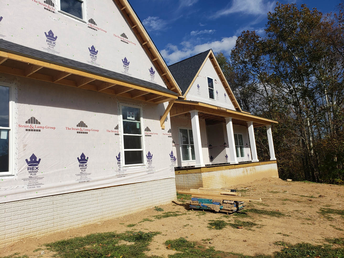 Framed house under construction with exposed plywood, several windows installed, pile of wood pallets in front, trees and cloudy sky in background