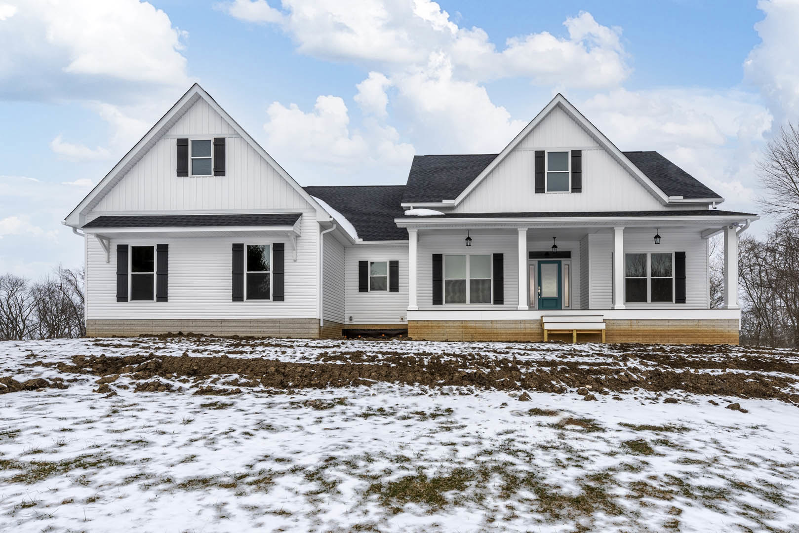 White house with black shutters and white-framed windows, snow-covered lawn with patches of grass, attached garage door, bare trees in the yard under a cloudy sky