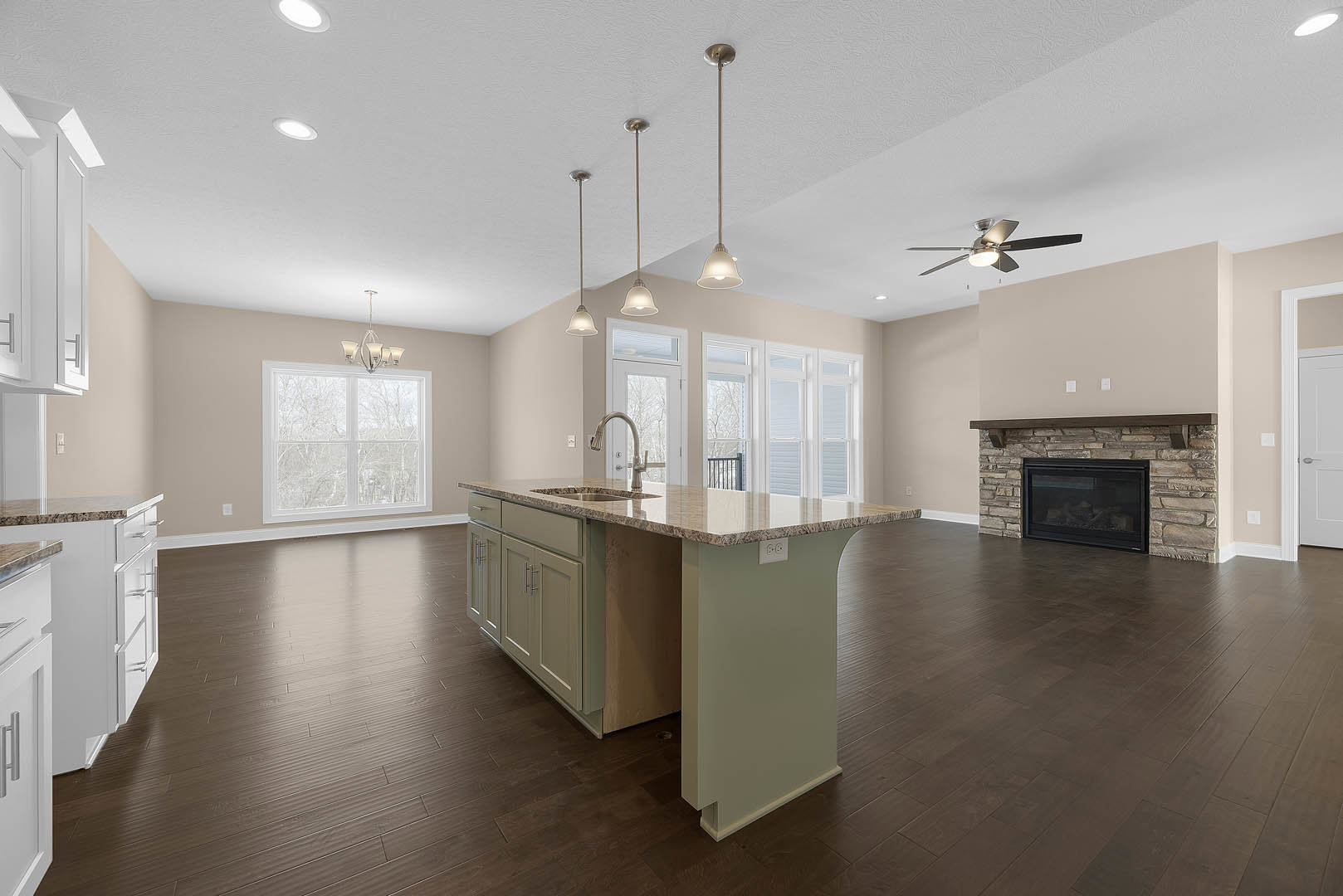 Open-concept kitchen and living room featuring a stone fireplace with glass doors, kitchen island with sink, wood cabinetry, pendant light fixtures, large window, and ceiling fan