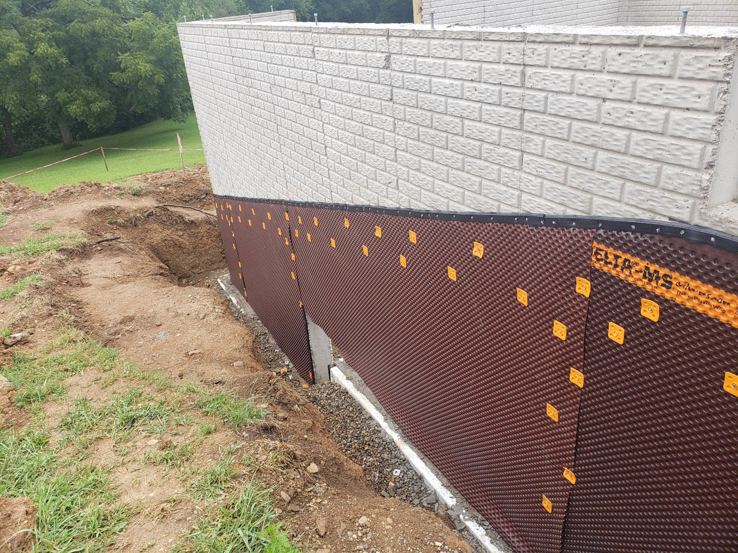 White brick exterior wall with black trim, brown and orange plastic panels, hole in the ground bordered by red rope, green grass field and trees in background