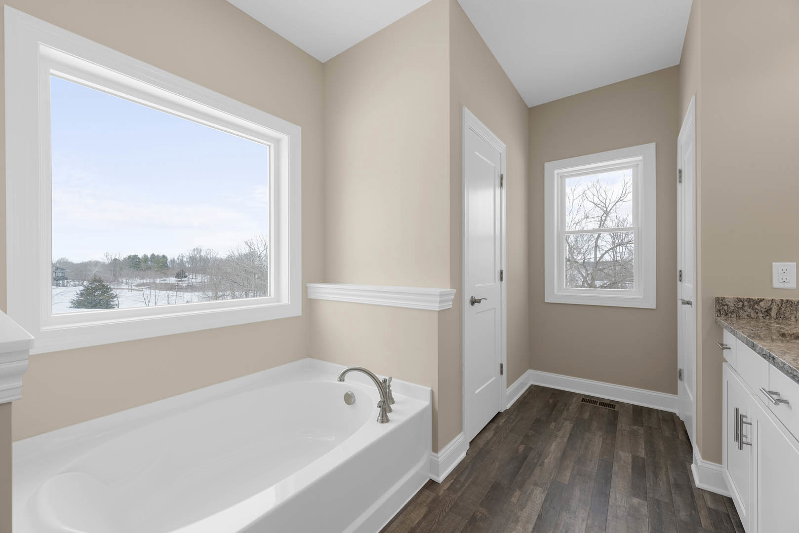 Freestanding white bathtub beneath large windows with snowy landscape views, chrome faucet, light tile flooring, and white door with silver handle