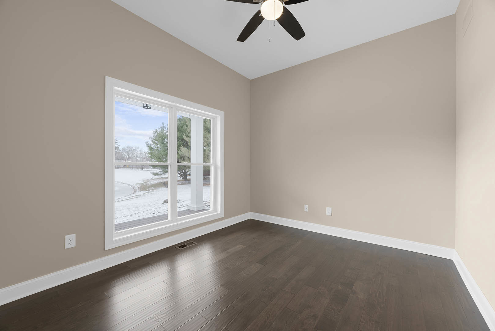 Hardwood floor room with white plaster walls, ceiling fan with light fixture, large window overlooking snowy yard and trees