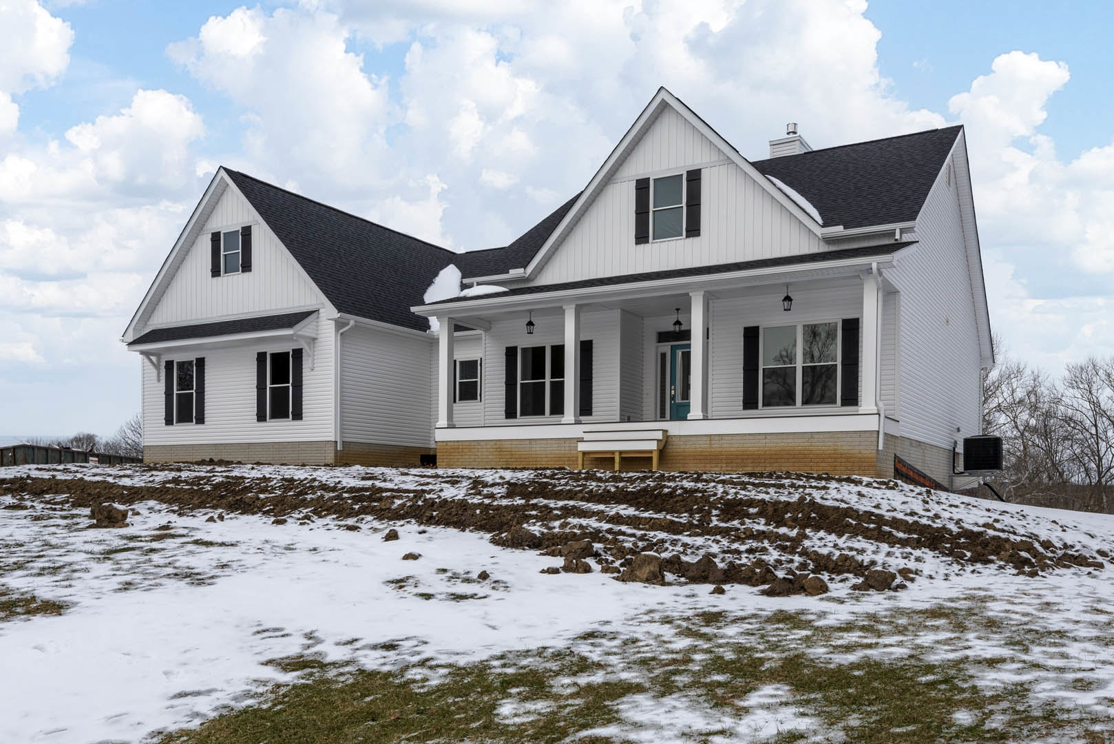 Two-story cottage with white siding, blue front door, snow covering the lawn, bare tree branches in the background, overcast sky