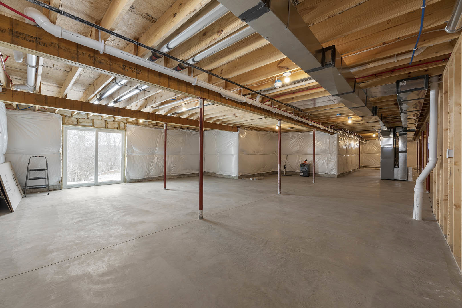 Spacious room with white walls, wooden beam ceiling, concrete floor, red support poles, four-step ladder, large window overlooking forest, exposed ceiling pipes