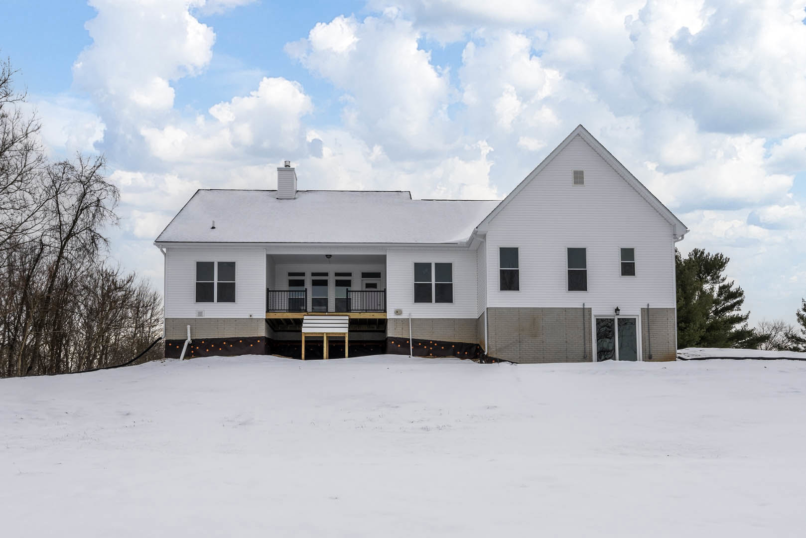 White house with black-framed windows, covered porch, and wooden deck, surrounded by snow-covered ground and a fence; bare trees and cloudy sky in the background.
