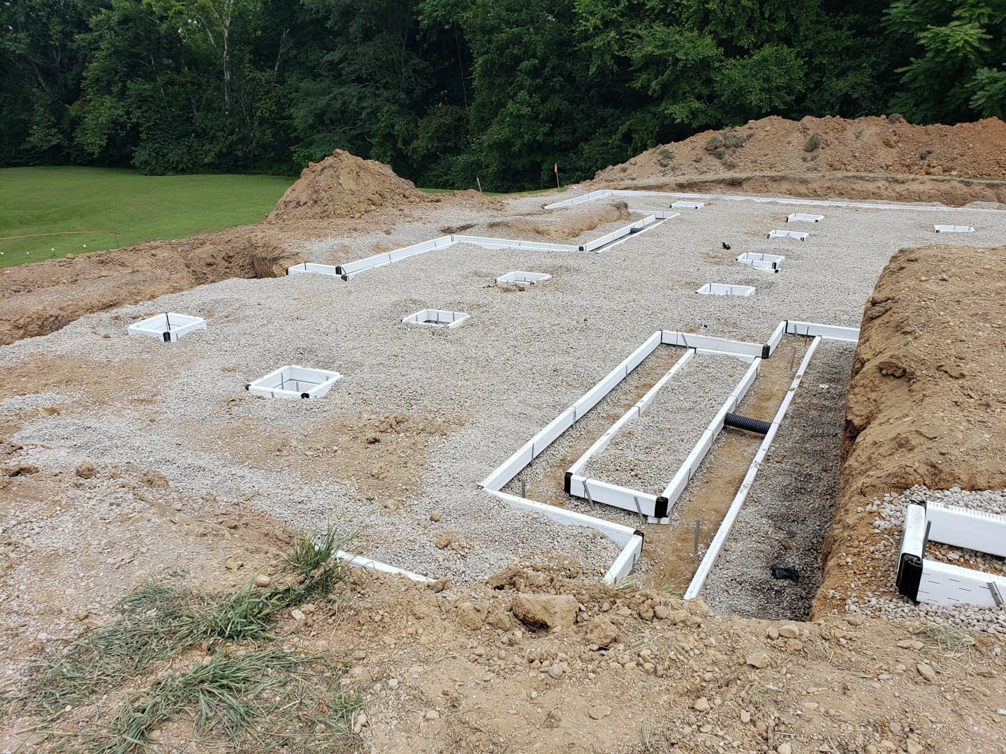 House foundation under construction with white pipes and plastic sheeting, metal rods embedded in a square white object, surrounded by soil and trees.