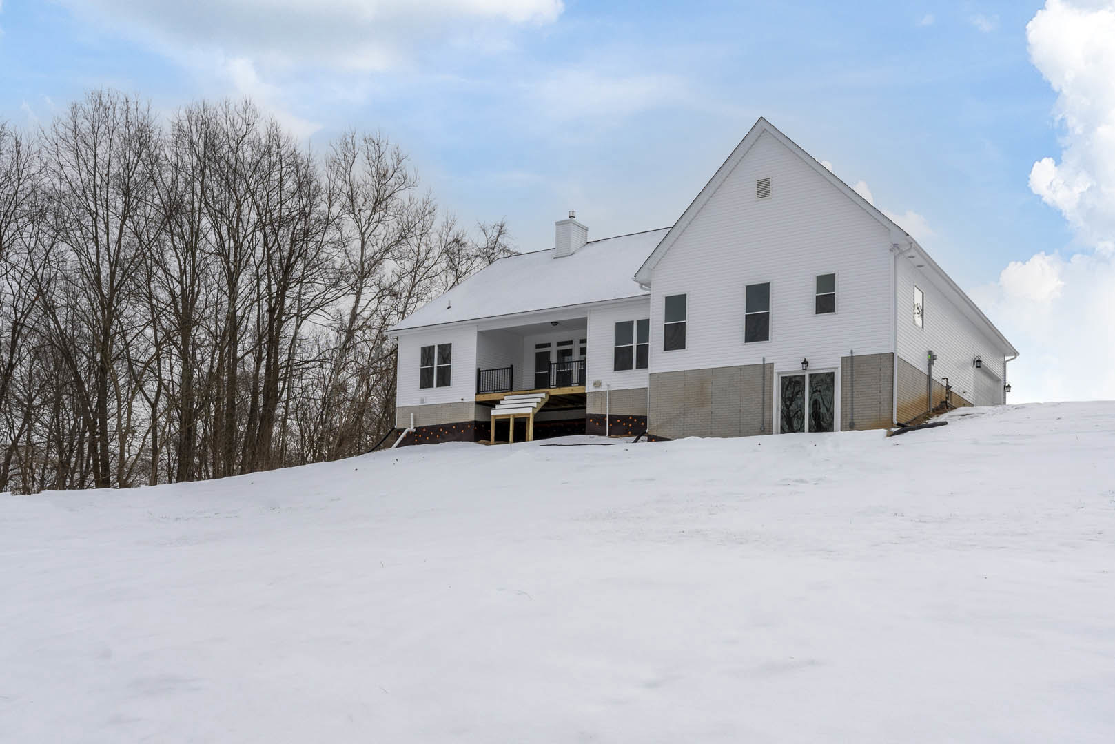 Two-story house with white siding, elevated deck, exterior staircase, and snow covering the ground and roof, surrounded by bare trees under a cloudy winter sky
