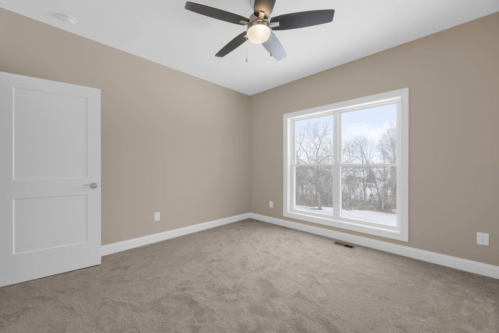 Carpeted room with white walls, ceiling fan with light, large window showing trees outside, white door with silver handle, white electrical outlet on wall, simple molding along