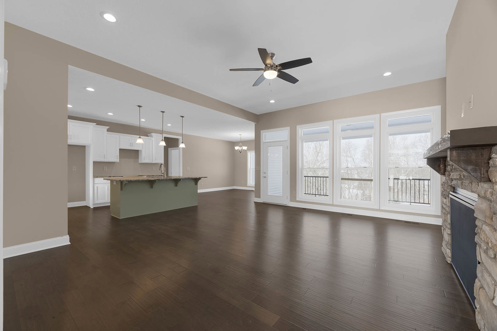 Spacious kitchen with dark wood flooring, central island featuring a sink, ceiling fan with light fixture, white-framed windows overlooking trees.