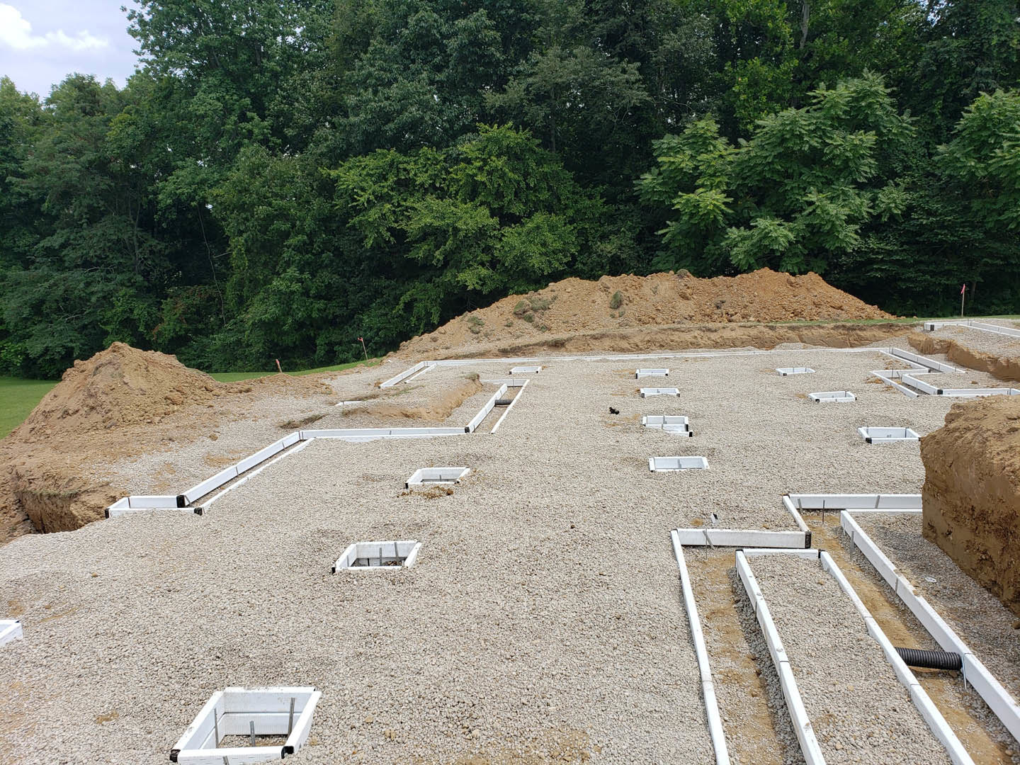 Dirt construction site bordered by trees, square concrete drain embedded in soil, excavation holes, and exposed white wall section