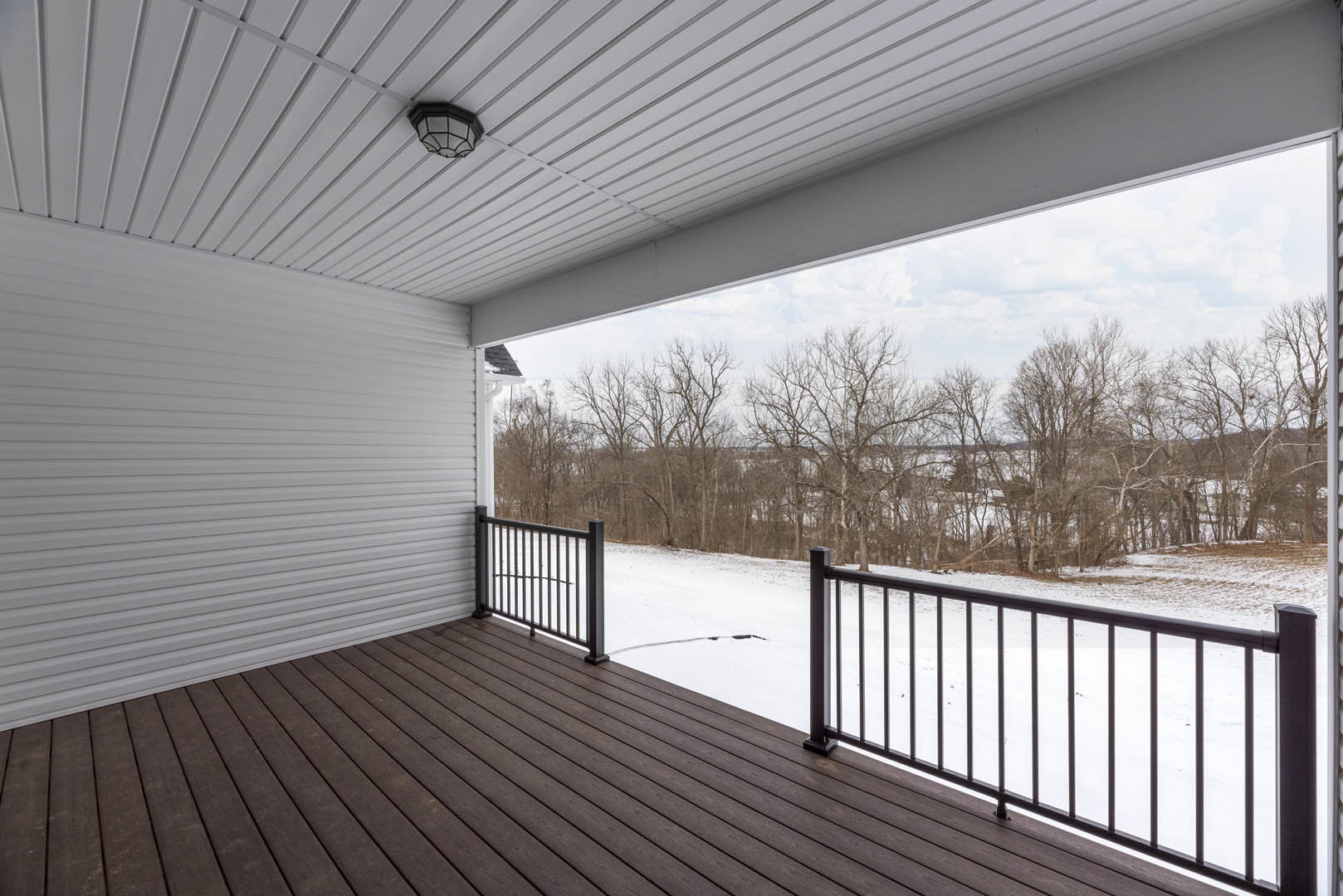 Wood deck with metal railing overlooking snowy yard, bare trees, and brick exterior wall with window