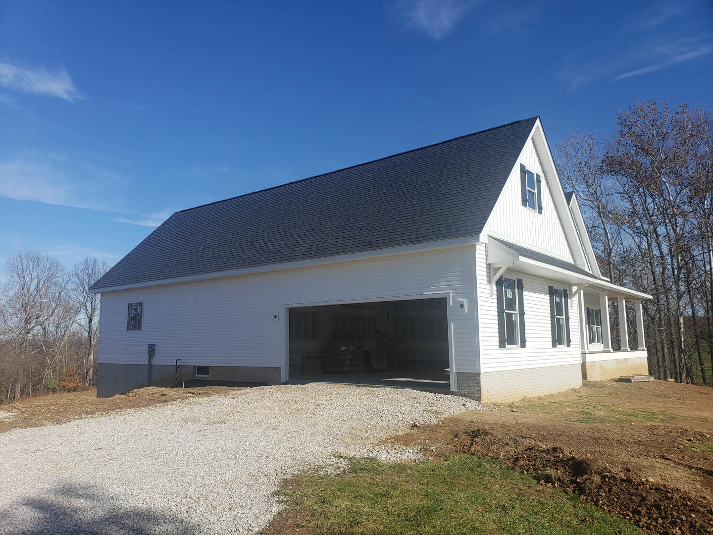 White two-story house with attached garage, gray shingle roof, leafless tree in front yard, gravel driveway, grassy lawn, and a window displaying a sign.