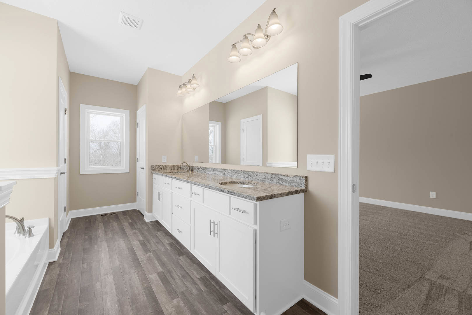 Bathroom featuring a marble countertop, expansive mirror, white bathtub with mesh cover, modern light fixtures, cabinetry, and window overlooking trees.