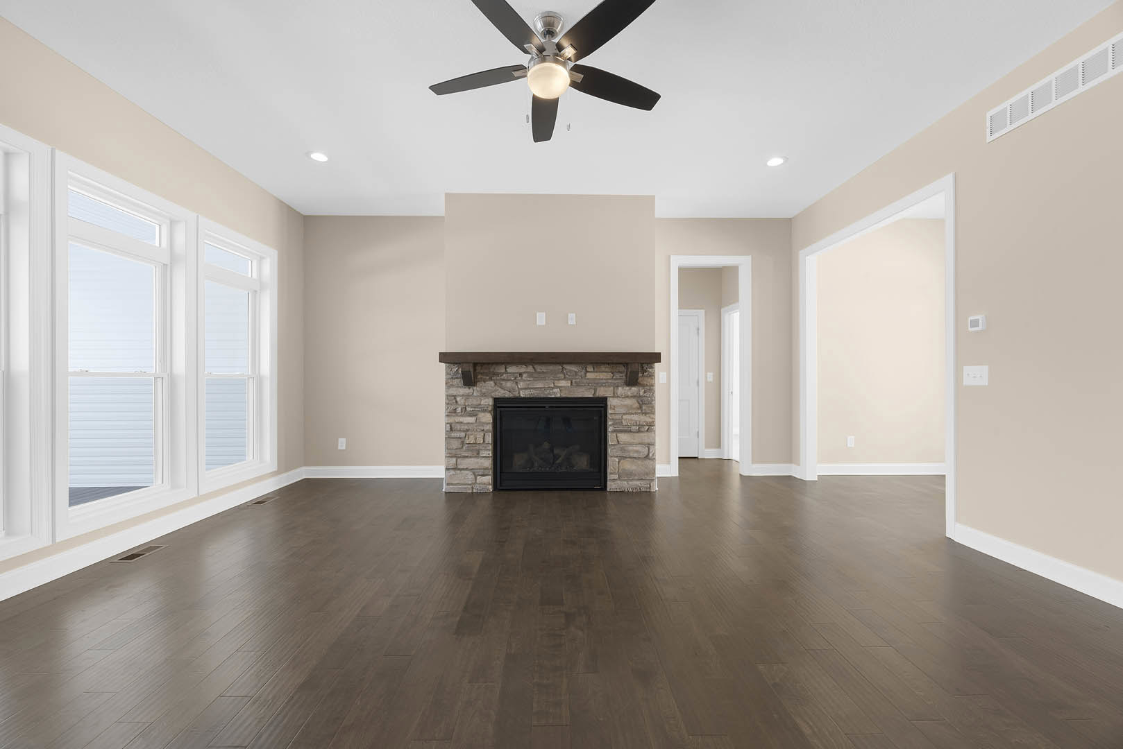 Living room with hardwood floors, stone fireplace with glass door, ceiling fan with light, white trim molding, window, and adjacent hallway with white door
