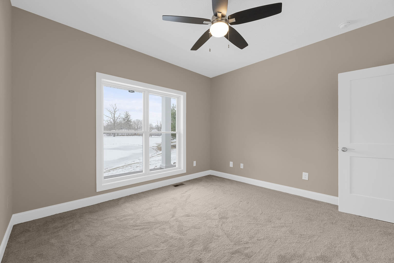 Carpeted room with white walls, ceiling fan with light fixture, large window overlooking snowy field, and paneled door