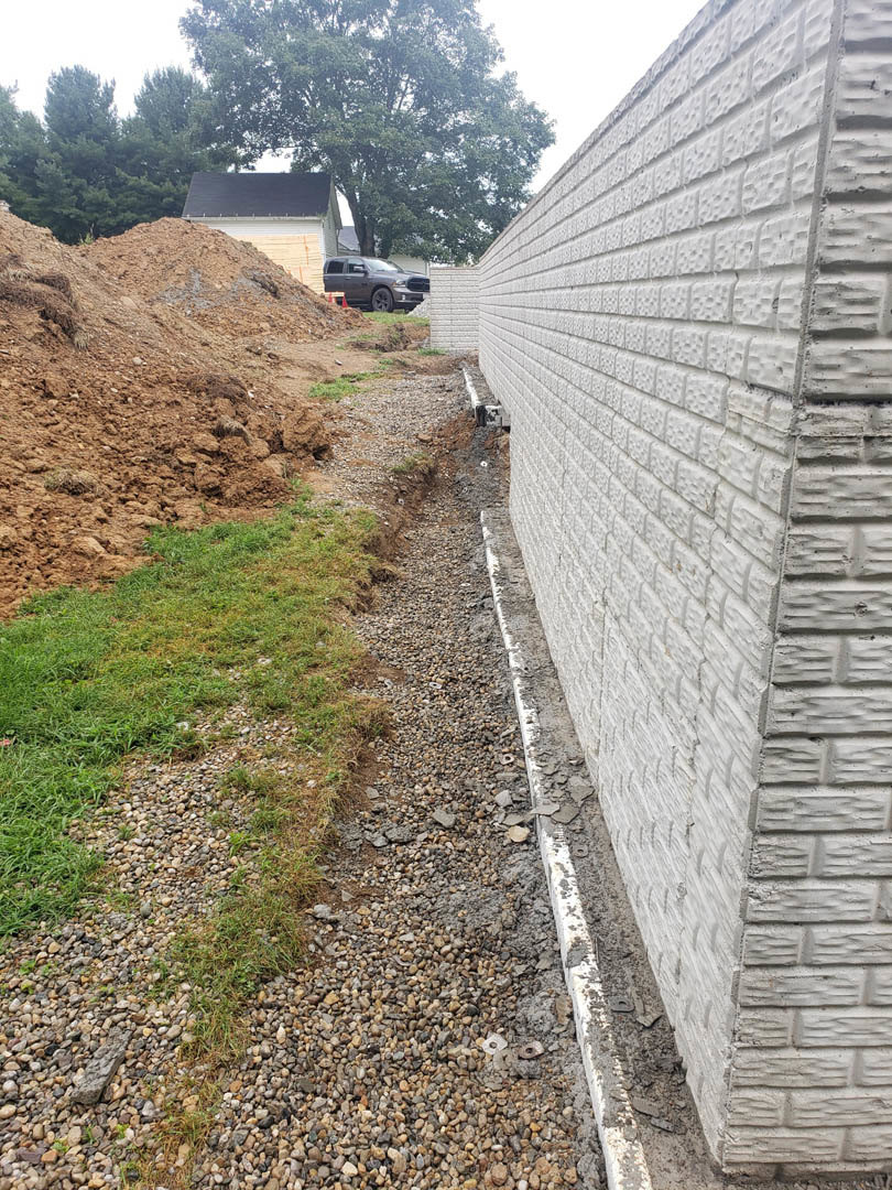 White brick exterior wall beside a dirt hill with gravel and grass, a car parked on the road, man standing in front of a pile of dirt, trees in the background